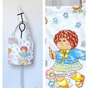 Pode incluir: Uma sacola branca com um padrão repetitivo de um personagem de desenho animado com cabelo ruivo encaracolado, vestindo uma jaqueta azul, laço amarelo e saia xadrez. O personagem segura um pincel, cercado por pequenas flores azuis e rosas.