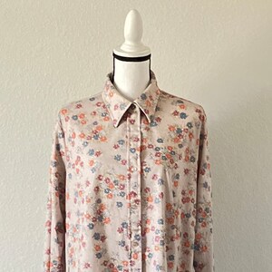Puede incluir: Una camisa beige de botones con estampado floral con peque&ntilde;as flores azules, rojas y naranjas. La camisa est&aacute; en un maniqu&iacute; blanco con una base de madera.