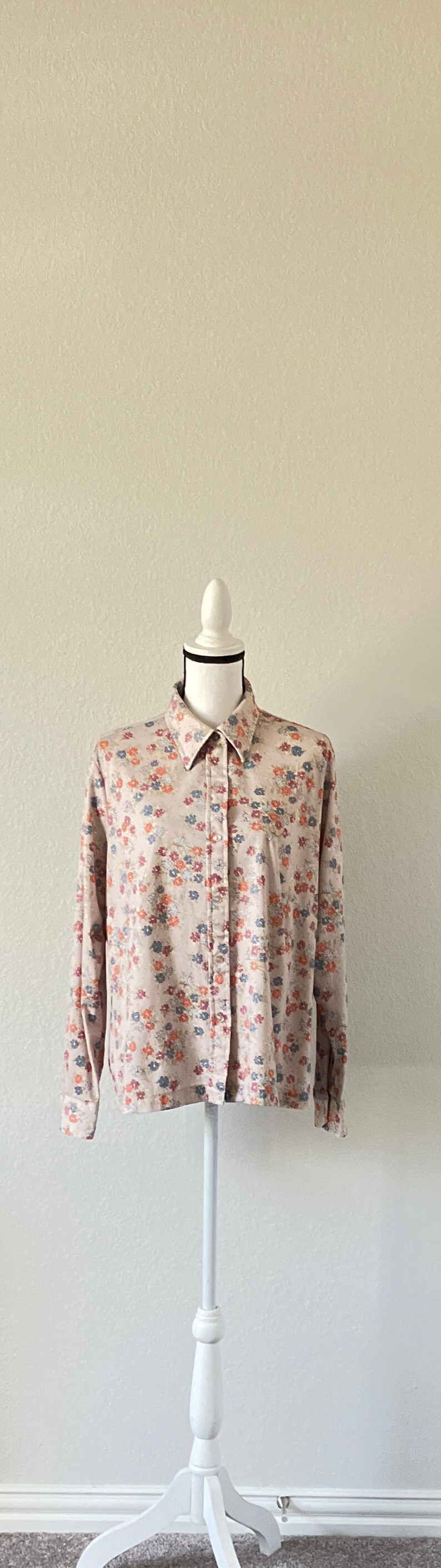 Puede incluir: Una camisa beige de botones con estampado floral con peque&ntilde;as flores azules, rojas y naranjas. La camisa est&aacute; en un maniqu&iacute; blanco con una base de madera.