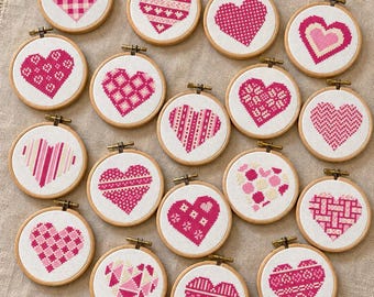 May include: A collection of sixteen small circular wooden embroidery hoops, each containing a unique cross-stitch heart design in various shades of pink, red, and white, arranged on a textured beige fabric surface.