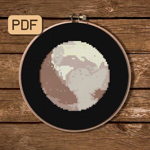 May include: A cross-stitch embroidery of a celestial body, possibly a planet or moon, in shades of beige, brown, and cream against a black background. The design is framed in a wooden hoop, with the text "PDF" in a tan box in the upper left corner.
