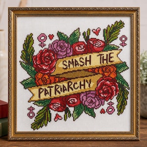 Puede incluir: Obra de arte de punto de cruz en un marco dorado. El diseño presenta una pancarta con las palabras "SMASH THE PATRIARCHY" rodeada de rosas rojas, moradas y naranjas, hojas verdes y símbolos de género. La obra está sobre un fondo blanco.
