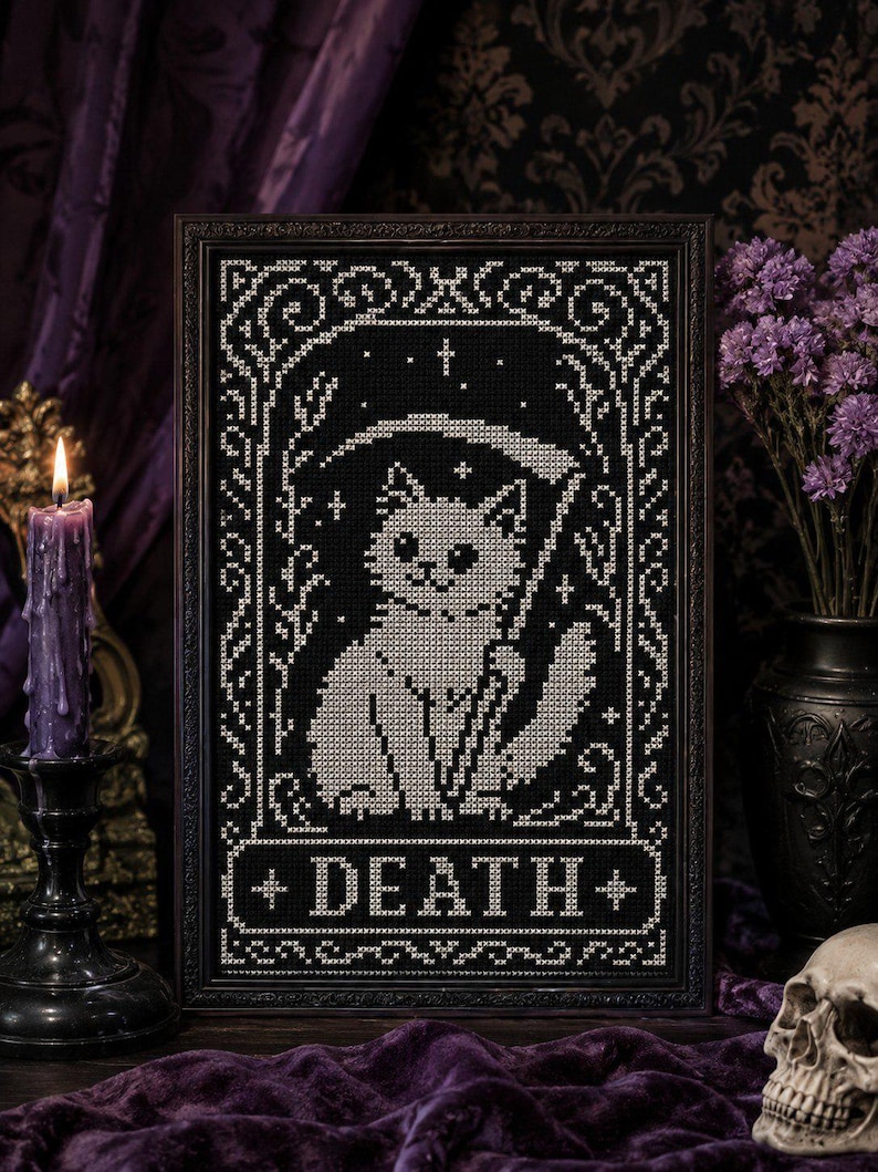 May include: A framed cross-stitch embroidery featuring a white cat holding a scythe, set against a black background. The design is enclosed in an ornate border with stars and includes the word DEATH in a rectangular banner at the bottom.