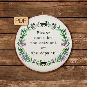 Könnte beinhalten: Ein weißer Stickrahmen mit einem floralen Kranzdesign und dem Text "Please don't let the cats out or the cops in". Es befinden sich zwei schwarze Katzensilhouetten im Design. Der Rahmen befindet sich auf einem hölzernen Hintergrund.