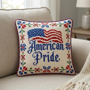 May include: A square, off-white pillow with a cross-stitched American flag and the words "American Pride" in blue. The pillow has a red and blue floral border and a red rope trim.