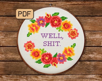May include: A white embroidery hoop with a floral wreath design and the text "WELL, SHIT." in purple letters. The hoop is on a wooden background.