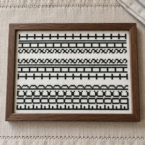 May include: A framed cross-stitch sampler featuring black geometric patterns on a white fabric background. The design consists of several horizontal rows of repeating linear motifs, including bars, squares, and scalloped edges, all enclosed in a dark wood frame.
