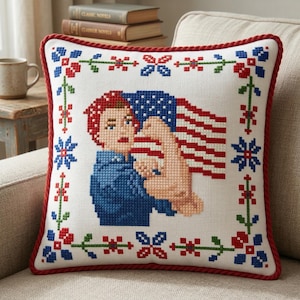 May include: Square decorative pillow with a cross-stitch design. The pillow features a central image of a person holding an American flag, surrounded by a border of red, white, and blue floral patterns. The pillow has a red rope trim.