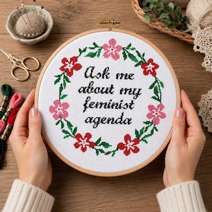Op de afbeelding: Een kruissteek borduurring met de tekst "Ask me about my feminist agenda" omringd door een bloemenkrans. De borduursels hebben rode en roze bloemen met groene bladeren. De ring wordt door twee handen vastgehouden.