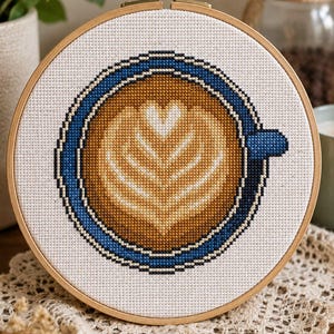Könnte beinhalten: Ein runder Stickrahmen aus Holz mit einem Kreuzstichmuster einer Kaffeetasse in der Draufsicht. Das Design zeigt eine blaue Tasse, gefüllt mit braunem Kaffee und einem weißen, herzförmigen Milchschaummuster in der Mitte des Getränks.