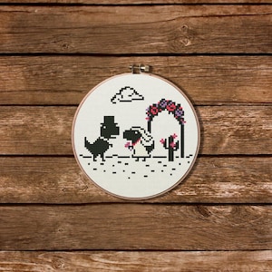 Op de afbeelding: Een kruissteekborduurwerk met een gepixeld dinosauruspaar dat trouwt. Het ontwerp omvat een bloemenboog, een wolk en een cactus, allemaal geborduurd op witte stof. De borduurring staat tegen een houten achtergrond.