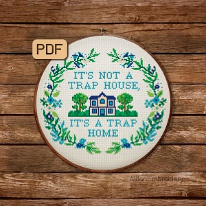 Puede incluir: Un aro de bordado blanco con un diseño de punto de cruz que dice "No es una casa trampa, es un hogar trampa". El diseño presenta una casa azul con ventanas amarillas y árboles verdes. El aro está rodeado de una corona floral verde y azul.