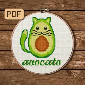 May include: Cross stitch pattern of an avocado shaped cat with the text "avocato" underneath. The pattern is in a white embroidery hoop on a wooden background.
