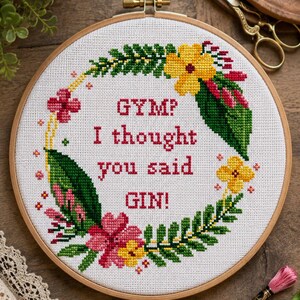 Könnte beinhalten: Ein runder Holzstickrahmen mit weißem Stoff und einem Kreuzstichmotiv. Der Text lautet 'GYM? I thought you said GIN!' in roten Buchstaben, umgeben von einem farbenfrohen Blumenkranz mit grünen Blättern sowie rosa und gelben Blüten.