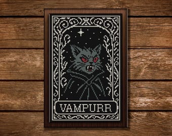 May include: Cross-stitch art of a gray cat with red eyes, dressed in a black cape, against a starry night background. The framed artwork features the word "VAMPURR" and decorative borders, set against a wooden backdrop.