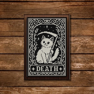 May include: A framed black and white cross-stitch featuring a white cat with a scythe, labelled "DEATH." The design includes decorative borders and stars on a black background. The frame is a dark brown colour.