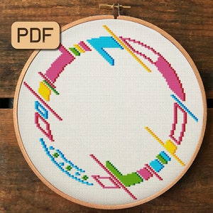 以下が含まれることがあります： 木製の刺繍枠に入ったクロスステッチのデザイン。ピンク、青、緑、黄色の幾何学的な形がカラフルに配置されています。「PDF」の文字が左上にあります。