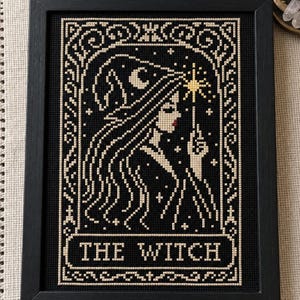 May include: A framed cross-stitch embroidery featuring a profile of a figure wearing a pointed hat, holding a star-tipped wand. The design uses black and cream thread with red lips and a yellow star, enclosed in a decorative border with the text THE WITCH.