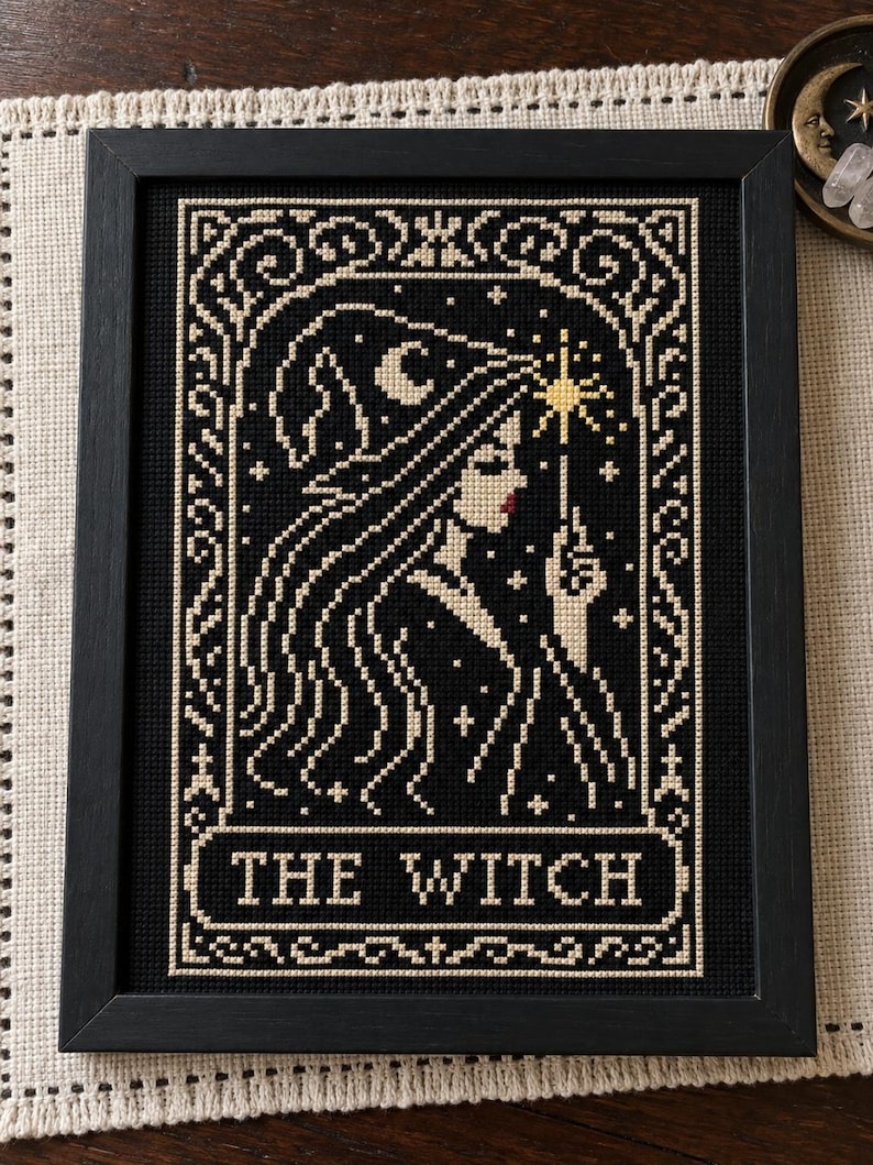 May include: A framed cross-stitch embroidery featuring a profile of a figure wearing a pointed hat, holding a star-tipped wand. The design uses black and cream thread with red lips and a yellow star, enclosed in a decorative border with the text THE WITCH.