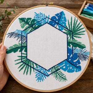 May include: A cross-stitch embroidery hoop featuring a tropical leaf design in shades of blue and green. The design includes a central hexagon shape, surrounded by various leaf patterns. The wooden hoop is held by a hand.