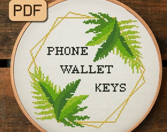 May include: Cross-stitch embroidery in a wooden hoop. The design features the words "PHONE WALLET KEYS" in black block letters, surrounded by green fern-like leaves and a gold geometric shape. A PDF icon is in the upper left corner.
