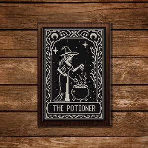 May include: A framed black and white cross-stitch depicting a witch stirring a cauldron, with the text "THE POTIONER" at the bottom. The artwork is set against a dark wooden background, creating a rustic and mystical aesthetic.