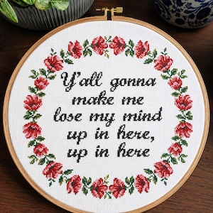 May include: A circular wooden embroidery hoop containing white fabric with a cross-stitched floral wreath of red flowers and green leaves surrounding the black text: Y'all gonna make me lose my mind up in here, up in here.