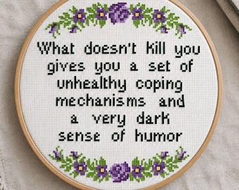 May include: A circular wooden embroidery hoop containing white fabric with a cross-stitched floral border in purple and green. The centre features black text reading: What doesn't kill you gives you a set of unhealthy coping mechanisms and a very dark sense of humour.
