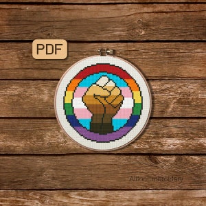 May include: A cross-stitch pattern of a raised fist in the center of a rainbow and transgender pride flag. The pattern is in a circular hoop and is ready to be stitched.