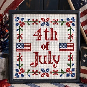 Op de afbeelding: Een vierkant ingelijst kruissteek borduurwerk op witte stof met de rode tekst 4th of July. Het ontwerp bevat twee kleine Amerikaanse vlaggen en een decoratieve rand van rode, witte, blauwe en groene bloemmotieven in een symmetrisch patroon.