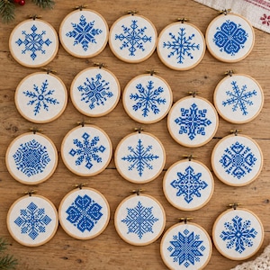 May include: A collection of nineteen circular wooden embroidery hoops, each containing white fabric with a unique blue cross-stitch snowflake pattern. The hoops are arranged in a grid, featuring various geometric snowflake designs in blue thread.