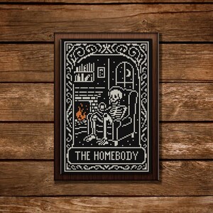 May include: A framed black and white cross-stitch depicting a skeleton relaxing in a chair by a fireplace. The text "THE HOMEBODY" is at the bottom of the frame. The frame is brown, and the background is a wooden surface.