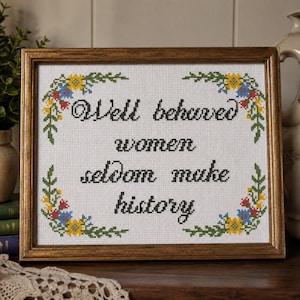 Puede incluir: Obra de arte en punto de cruz enmarcada con la cita "Well behaved women seldom make history". El texto está en escritura negra sobre un fondo blanco, con diseños florales en las esquinas en amarillo, azul y rojo. El marco es marrón cálido.