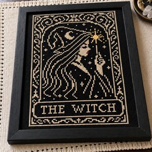 May include: A framed cross-stitch embroidery featuring a profile of a figure wearing a pointed hat and holding a star-tipped wand. The design is stitched in cream thread on a black background, with the text THE WITCH centered at the bottom.