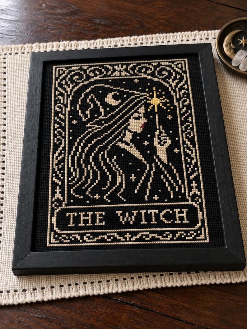 May include: A framed cross-stitch embroidery featuring a profile of a figure wearing a pointed hat and holding a star-tipped wand. The design is stitched in cream thread on a black background, with the text THE WITCH centered at the bottom.