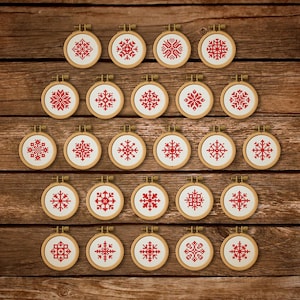 Puede incluir: Una colección de veintiún pequeños aros de bordado con diseños de copos de nieve rojos sobre un fondo blanco. Los aros están dispuestos sobre una superficie de madera, mostrando varios patrones de copos de nieve. Los aros de madera tienen aproximadamente 5 cm de diámetro.