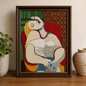May include: Framed cross-stitch artwork depicting a seated figure in a colorful, abstract style. The piece features a woman with blonde hair, wearing a white top, and seated in a red chair. The artwork is framed in a dark wood frame.