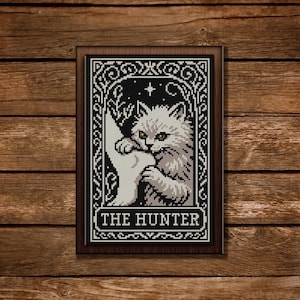 Puede incluir: Arte de punto de cruz enmarcado que presenta un gato blanco sosteniendo una luna creciente contra un cielo nocturno estrellado. La obra está bordeada por diseños ornamentados en blanco y negro y el texto "THE HUNTER".