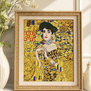 May include: A framed diamond painting depicting a portrait of a person with dark hair, wearing a high-collared garment. The artwork features a vibrant gold, yellow, and black mosaic pattern with geometric shapes and intricate details in a gold frame.