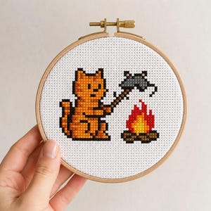 May include: A circular wooden embroidery hoop containing white Aida fabric with a pixel art cross-stitch design. The design features an orange cat sitting by a small campfire, holding a stick with a grey mouse roasted over the flames.