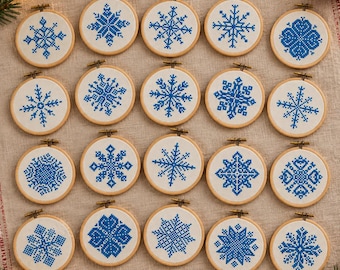 May include: Twenty circular wooden embroidery hoops arranged in a grid, each containing white fabric with a unique blue cross-stitch snowflake pattern. The hoops feature a small metal tightening screw at the top of each frame.