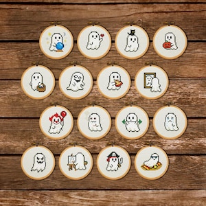 May include: Sixteen small, circular embroidery hoops, each displaying a unique pixelated ghost design. The ghosts are shown with different accessories and expressions, set against a wooden background. The hoops are framed in natural wood.