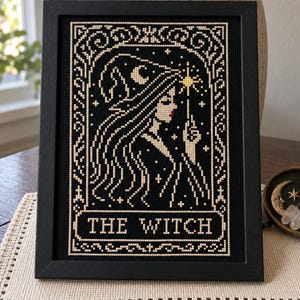 May include: A framed cross-stitch embroidery featuring a profile of a person wearing a pointed hat, holding a star-tipped wand. The design is stitched in cream thread on a black background with the text THE WITCH centered at the bottom.