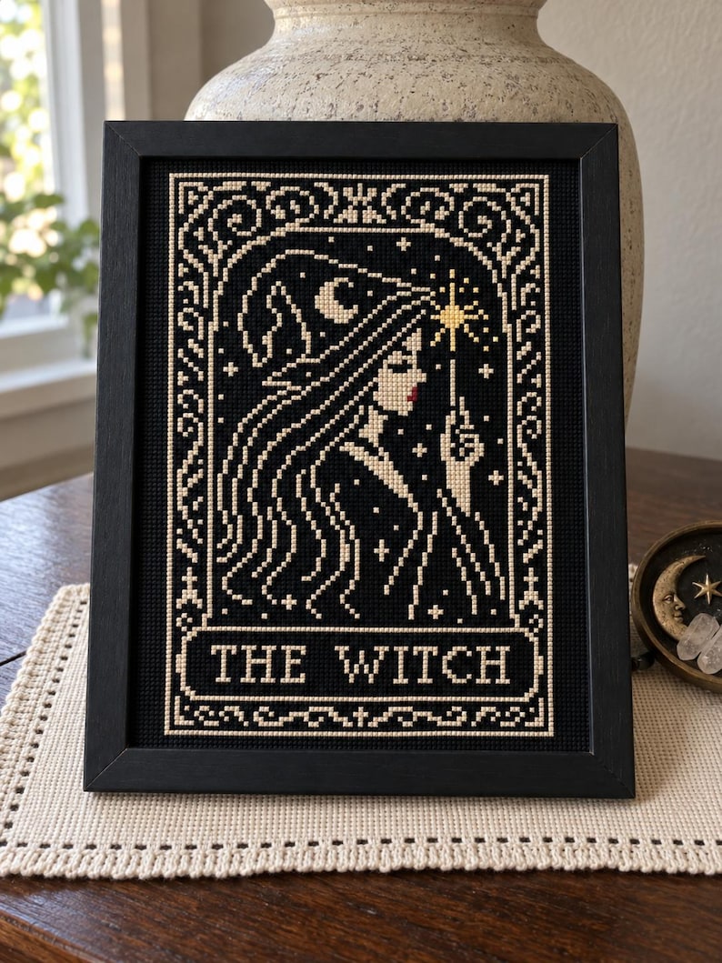 May include: A framed cross-stitch embroidery featuring a profile of a person wearing a pointed hat, holding a star-tipped wand. The design is stitched in cream thread on a black background with the text THE WITCH centered at the bottom.