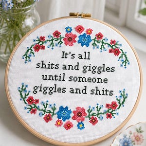 May include: A circular wooden embroidery hoop containing white fabric with a cross-stitch design. The text reads, "It's all shits and giggles until someone giggles and shits," surrounded by a colourful floral wreath of red, blue, and green thread.