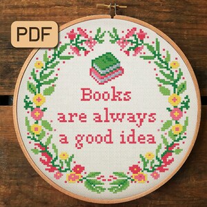 May include: A white embroidery hoop with a floral wreath design and the text "Books are always a good idea". The wreath is made of green leaves, pink and yellow flowers, and a stack of green, blue, and pink books.