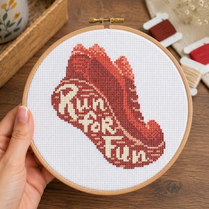 May include: A cross-stitch embroidery hoop featuring a red running shoe design with the words "Run for Fun" in cream-colored thread. The embroidery is set against a white background and framed by a wooden hoop.