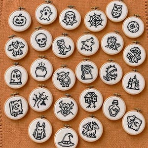 May include: A collection of twenty-six small circular wooden embroidery hoops, each containing a white fabric square with a black pixel-art cross-stitch design featuring various spooky, fantasy, and Halloween-themed icons.