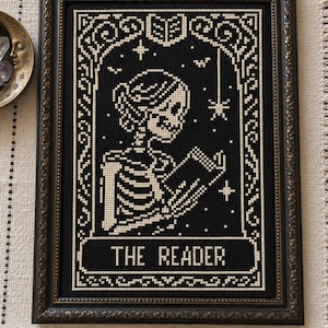 Könnte beinhalten: Ein gerahmtes Kreuzstich-Stickbild auf schwarzem Stoff, das ein weißes Skelett beim Lesen eines offenen Buches zeigt. Das Design enthält Sterne, eine Spinne und ein offenes Buchsymbol oben sowie den Text THE READER unten.