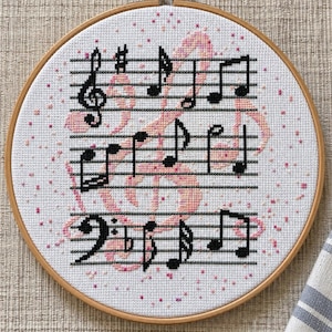May include: A circular wooden embroidery hoop containing white fabric with a cross-stitch design. The pattern features black musical notes and staves overlaid on a large, pale pink treble clef, accented by small pink confetti-like stitches.